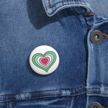 Close-up of a white circular pride button featuring a nested heart design inspired by the Abrosexual pride flag in greens, white, and pinks, attached to a blue denim jacket at the right chest with the button clearly visible.