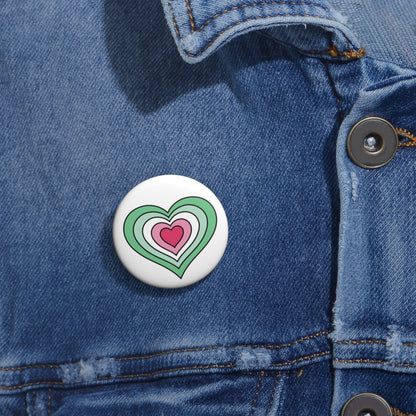 Close-up of a white circular pride button featuring a nested heart design inspired by the Abrosexual pride flag in greens, white, and pinks, attached to a blue denim jacket at the right chest with the button clearly visible.