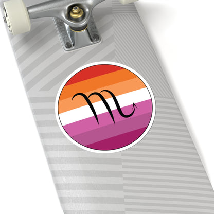 Close-up of the bottom right corner of a silver laptop featuring a 6 inch by 6 inch circular zodiac sticker with a centered Scorpio glyph on a Lesbian pride flag style background applied to the surface.