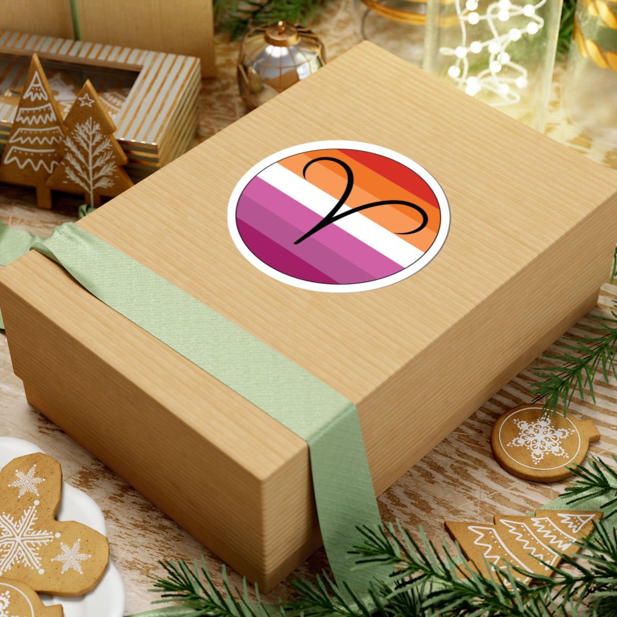 Gift box with a green ribbon on a festive background with cookies and Christmas decorations. Aires Lesbian pride sticker centered on the box.