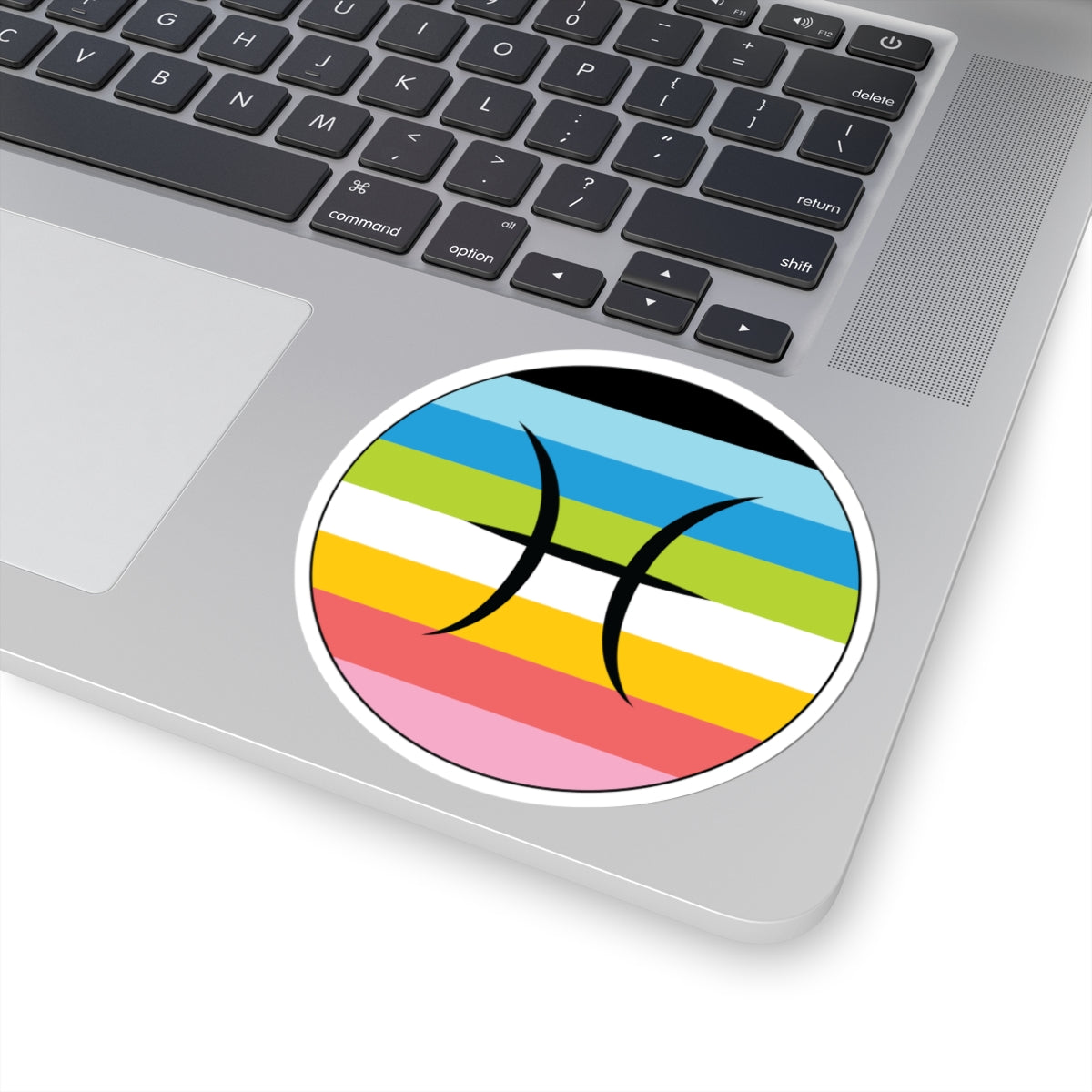 Close-up of the bottom right corner of a silver laptop featuring a 4 inch by 4 inch circular zodiac sticker with a centered Pisces glyph on a Queer pride flag style background applied to the surface.