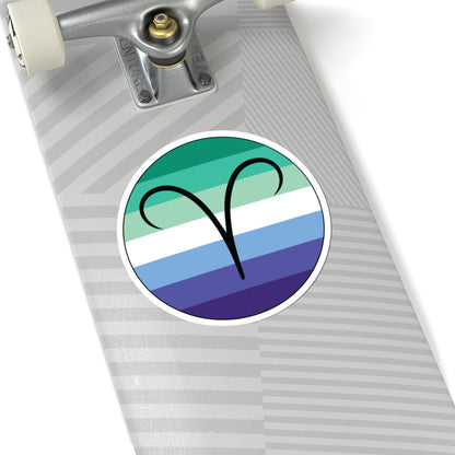 Close-up of the bottom right corner of a silver laptop featuring a 6 inch by 6 inch circular zodiac sticker with a centered Aires glyph on a Gay Men pride flag style background applied to the surface.