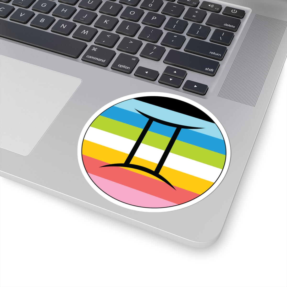 Close-up of the bottom right corner of a silver laptop featuring a 4 inch by 4 inch circular zodiac sticker with a centered Gemini glyph on a Queer pride flag style background applied to the surface.