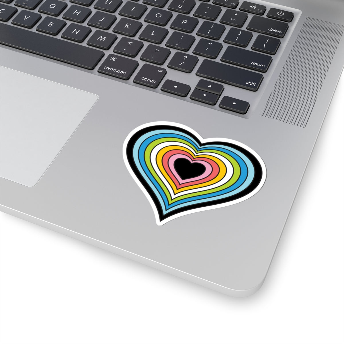 Close-up of the bottom right corner of a silver laptop featuring a 3 inch by 3 inch pride flag heart sticker in a Queer flag style applied to the surface.