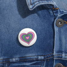 Close-up of a white circular pride button featuring a nested heart design inspired by the Trigender pride flag in pink, purple and green, attached to a blue denim jacket at the right chest with the button clearly visible.