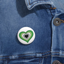Close-up of a white circular pride button featuring a nested heart design inspired by the Aromantic pride flag in greens, white gray and black, attached to a blue denim jacket at the right chest with the button clearly visible.