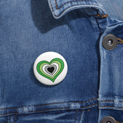 Close-up of a white circular pride button featuring a nested heart design inspired by the Aromantic pride flag in greens, white gray and black, attached to a blue denim jacket at the right chest with the button clearly visible.