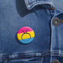Close-up of a white circular pride button featuring the Taurus zodiac glyph centered over a Pansexual pride flag background in pink, yellow, and blue, attached to a blue denim jacket at the right chest with the button clearly visible.
