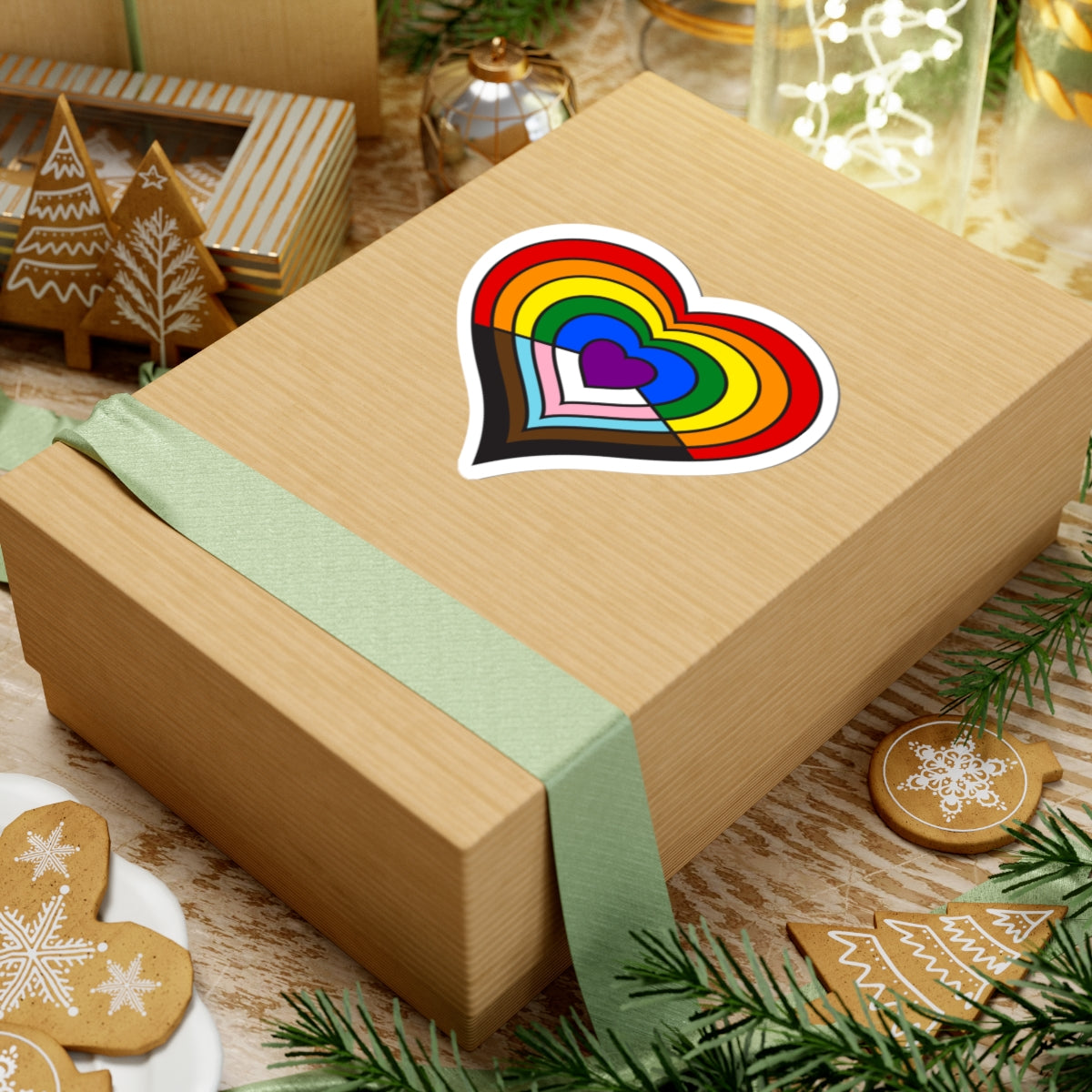 Gift box with a green ribbon on a festive background with cookies and Christmas decorations. Heart-shaped progress pride sticker centered on the box.