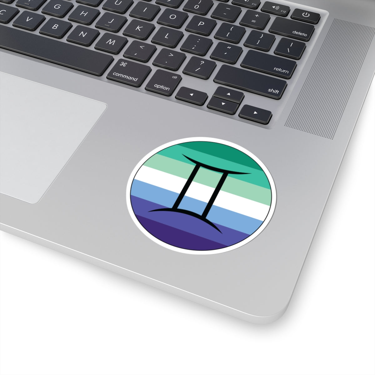 Close-up of the bottom right corner of a silver laptop featuring a 3 inch by 3 inch circular zodiac sticker with a centered Gemini glyph on a Gay Men pride flag style background applied to the surface.