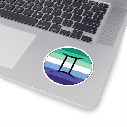 Close-up of the bottom right corner of a silver laptop featuring a 3 inch by 3 inch circular zodiac sticker with a centered Gemini glyph on a Gay Men pride flag style background applied to the surface.