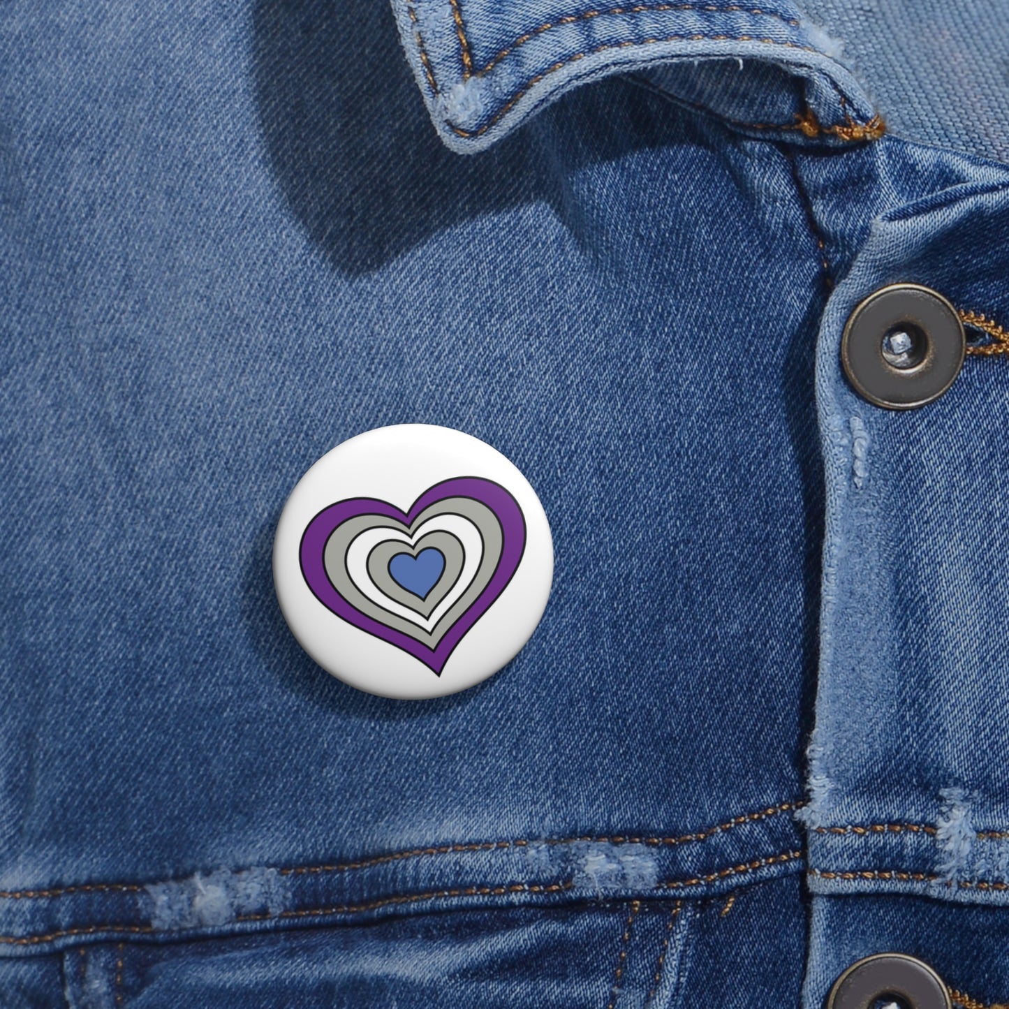 Close-up of a white circular pride button featuring a nested heart design inspired by the Greysexual pride flag in purple, gray, white and blue, attached to a blue denim jacket at the right chest with the button clearly visible.