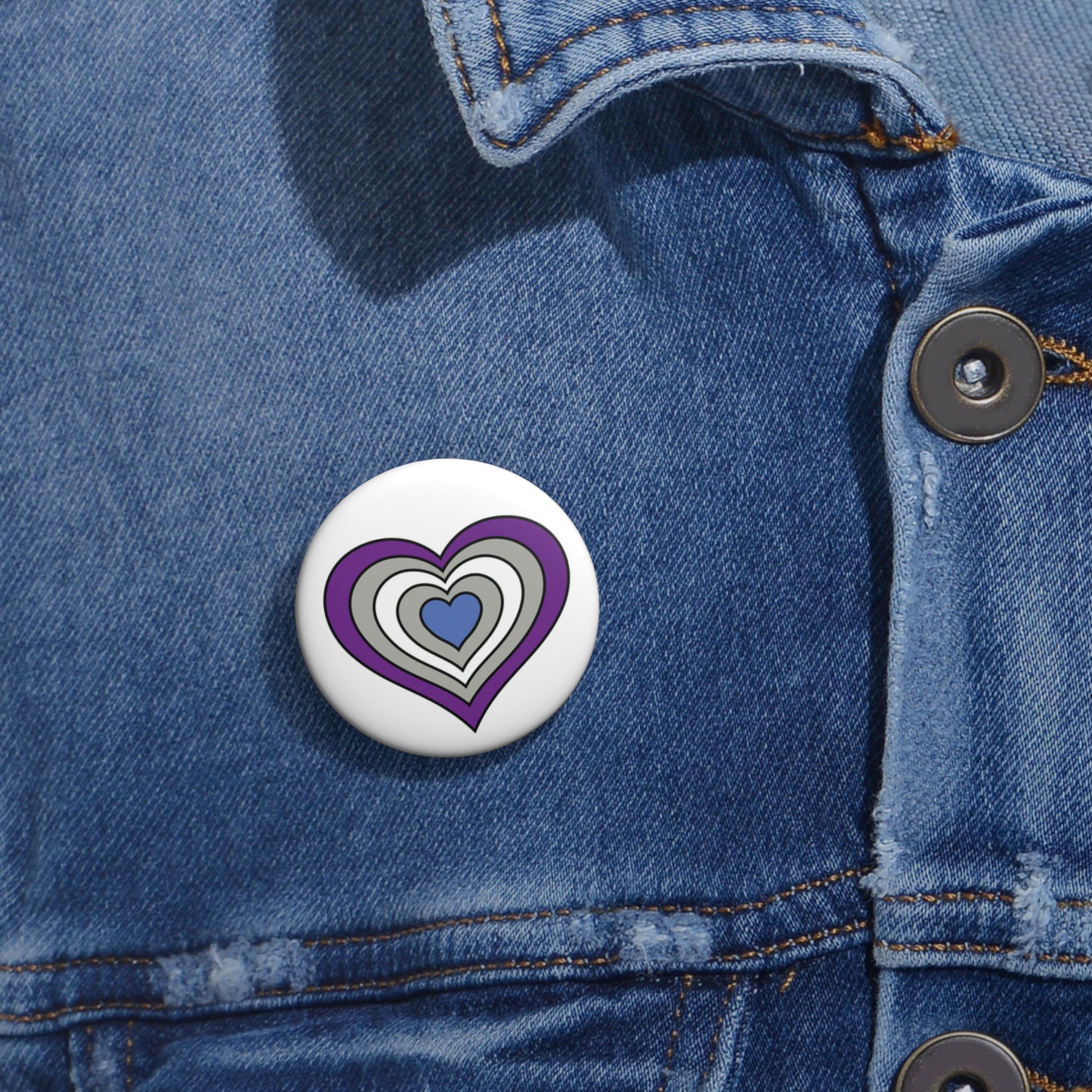 Close-up of a white circular pride button featuring a nested heart design inspired by the Greysexual pride flag in purple, gray, white and blue, attached to a blue denim jacket at the right chest with the button clearly visible.