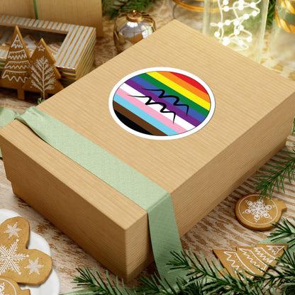 Gift box with a green ribbon on a festive background with cookies and Christmas decorations. Aquarius Progress pride sticker centered on the box.