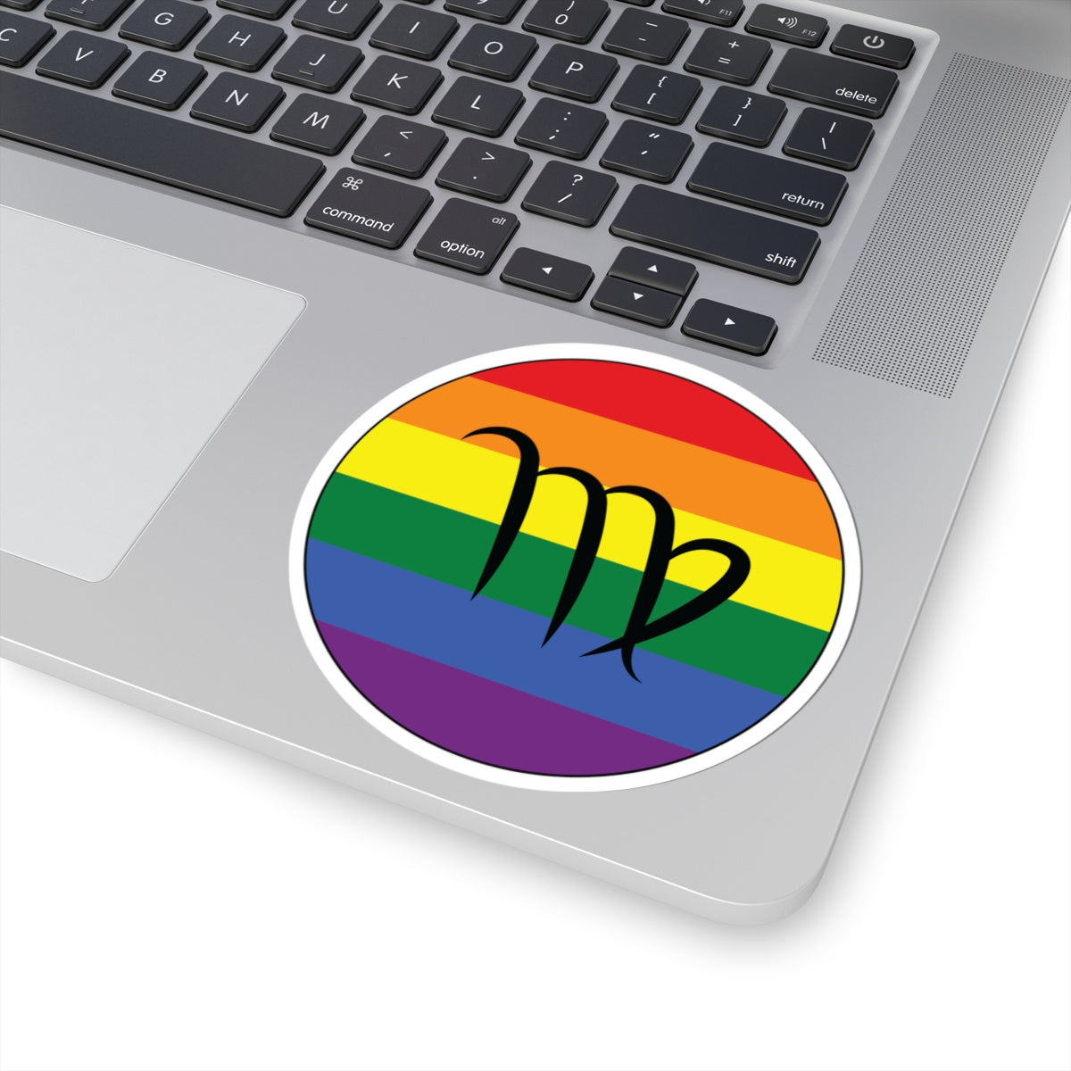 Close-up of the bottom right corner of a silver laptop featuring a 4 inch by 4 inch circular zodiac sticker with a centered Virgo glyph on a Rainbow pride flag style background applied to the surface.