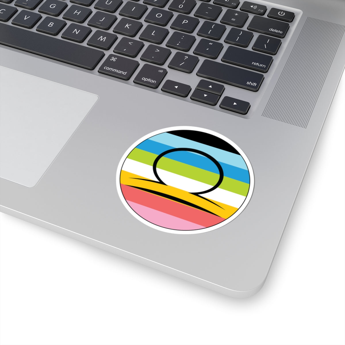 Close-up of the bottom right corner of a silver laptop featuring a 3 inch by 3 inch circular zodiac sticker with a centered Libra glyph on a Queer pride flag style background applied to the surface.