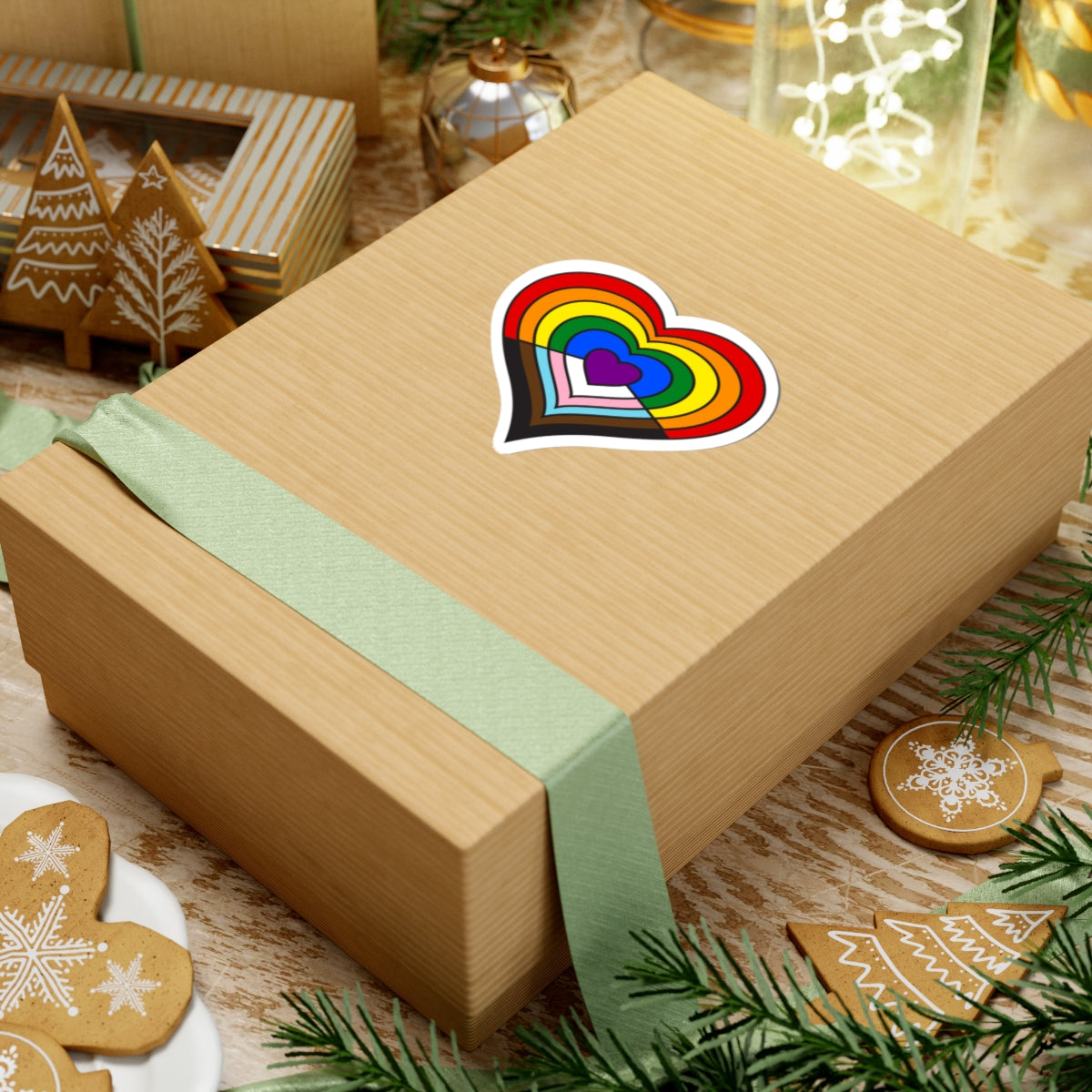Gift box with a green ribbon on a festive background with cookies and Christmas decorations. Heart-shaped progress pride sticker centered on the box.