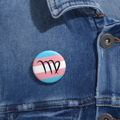 Close-up of a white circular pride button featuring the Virgo zodiac glyph centered over a transgender pride flag background in light blue, pink, and white, attached to a blue denim jacket at the right chest with the button clearly visible.