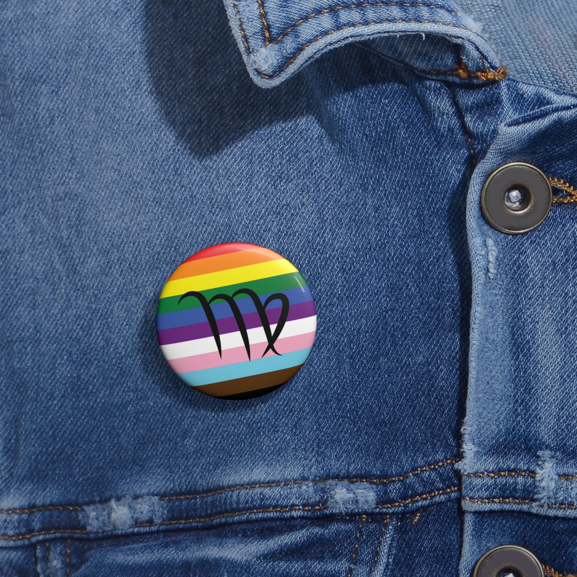 Close-up of a white circular pride button featuring the Virgo zodiac glyph centered over a Progress Pride flag background in rainbow stripes with black, brown, light blue, pink, and white elements, attached to a blue denim jacket at the right chest with the button clearly visible.