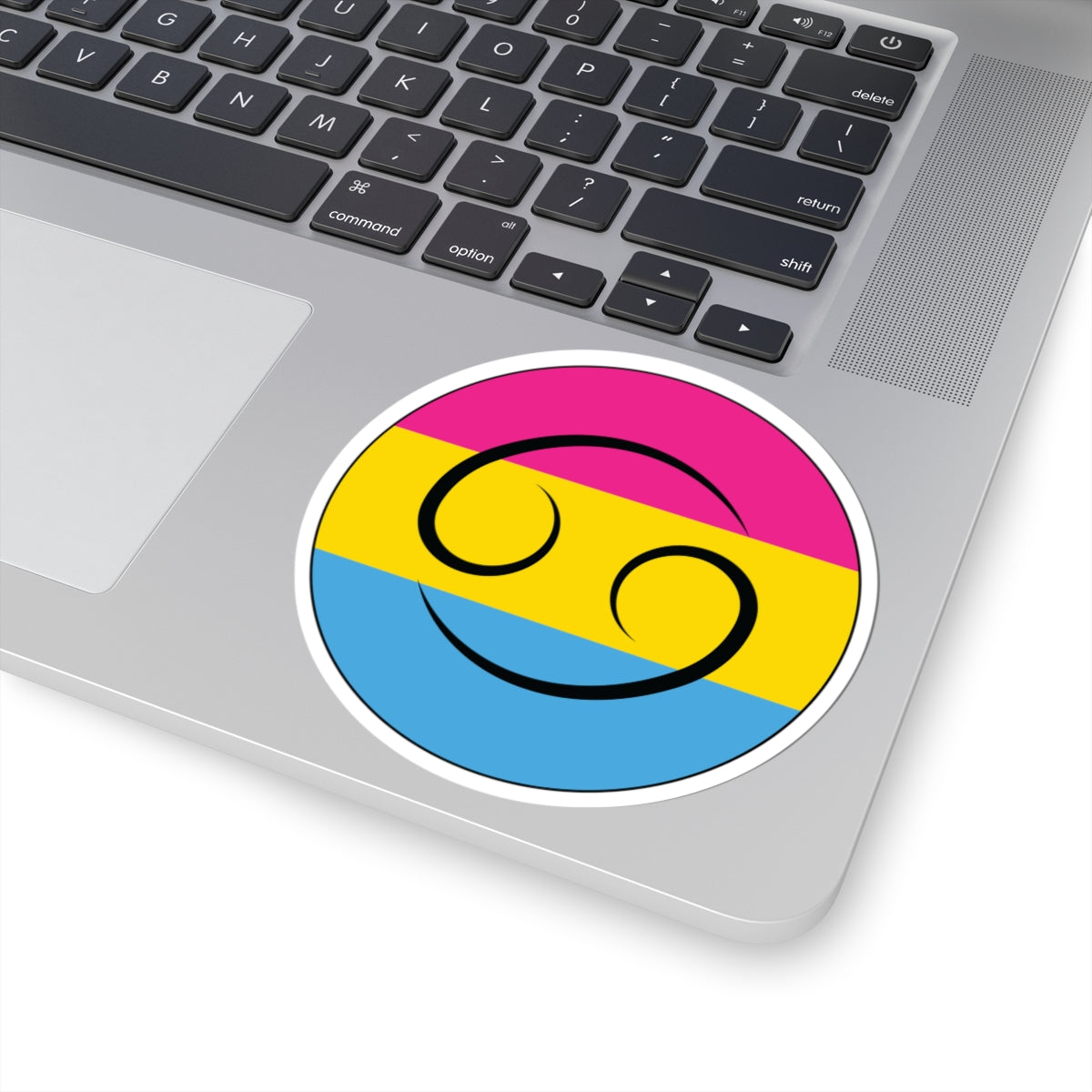 Close-up of the bottom right corner of a silver laptop featuring a 4 inch by 4 inch circular zodiac sticker with a centered Cancer glyph on a Pansexual pride flag style background applied to the surface.