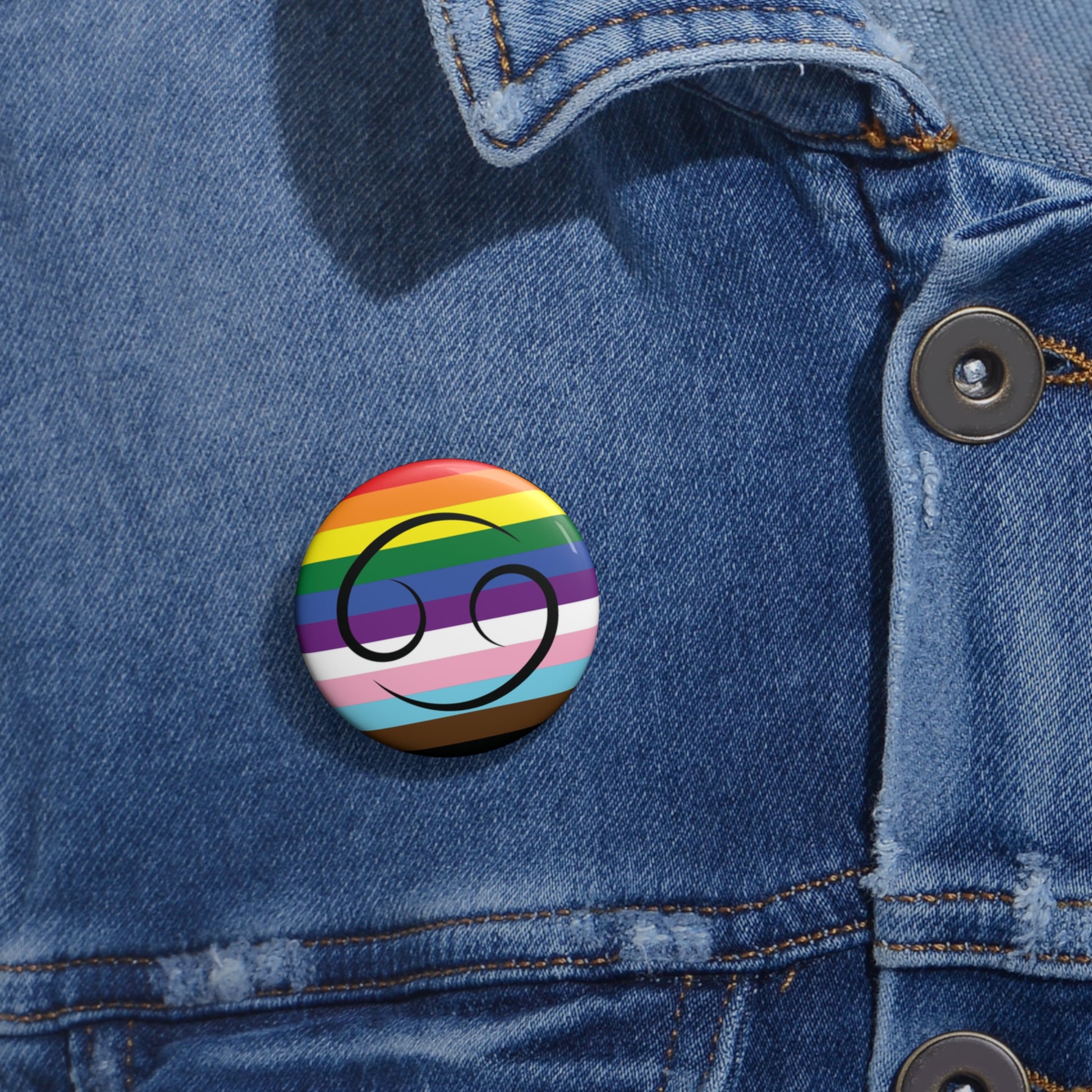 Close-up of a white circular pride button featuring the Cancer zodiac glyph centered over a Progress Pride flag background in rainbow stripes with black, brown, light blue, pink, and white elements, attached to a blue denim jacket at the right chest with the button clearly visible.