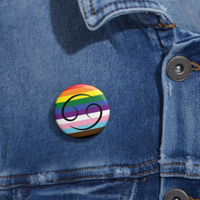 Close-up of a white circular pride button featuring the Cancer zodiac glyph centered over a Progress Pride flag background in rainbow stripes with black, brown, light blue, pink, and white elements, attached to a blue denim jacket at the right chest with the button clearly visible.