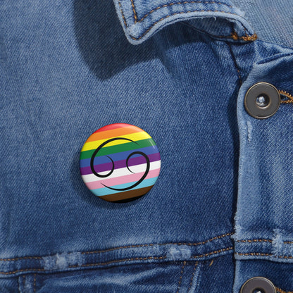 Close-up of a white circular pride button featuring the Cancer zodiac glyph centered over a Progress Pride flag background in rainbow stripes with black, brown, light blue, pink, and white elements, attached to a blue denim jacket at the right chest with the button clearly visible.