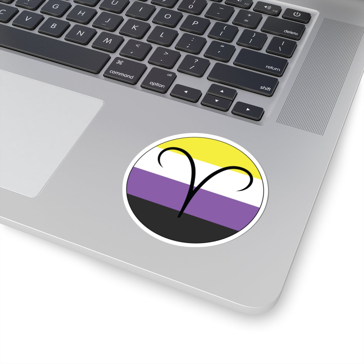 Close-up of the bottom right corner of a silver laptop featuring a 3 inch by 3 inch circular zodiac sticker with a centered Aires glyph on a Nonbinary pride flag style background applied to the surface.