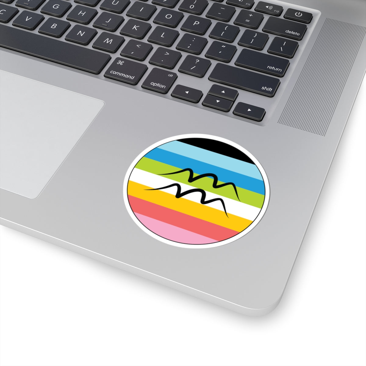 Close-up of the bottom right corner of a silver laptop featuring a 3 inch by 3 inch circular zodiac sticker with a centered Aquarius glyph on a Queer pride flag style background applied to the surface.