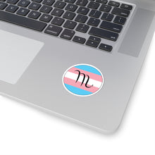 Close-up of the bottom right corner of a silver laptop featuring a 2 inch by 2 inch circular zodiac sticker with a centered Scorpio glyph on a Transgender pride flag style background applied to the surface.