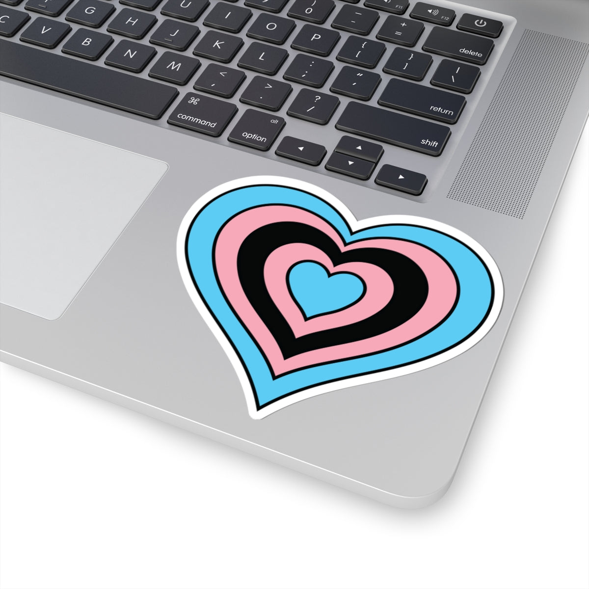 Close-up of the bottom right corner of a silver laptop featuring a 4 inch by 4 inch pride flag heart sticker in a Black Trans flag style applied to the surface.