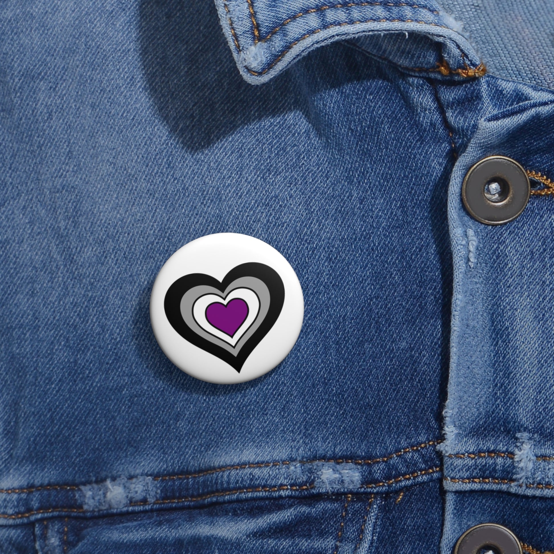 Close-up of a white circular pride button featuring a nested heart design inspired by the Asexual pride flag in black, gray, white and purple, attached to a blue denim jacket at the right chest with the button clearly visible.