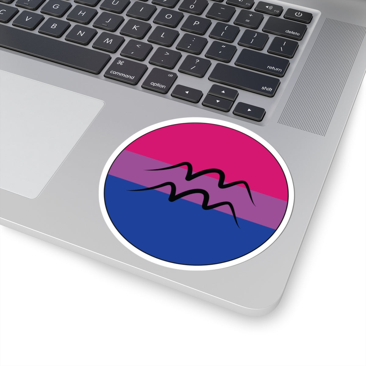Close-up of the bottom right corner of a silver laptop featuring a 4 inch by 4 inch circular zodiac sticker with a centered Aquarius glyph on a Bisexual pride flag style background applied to the surface.