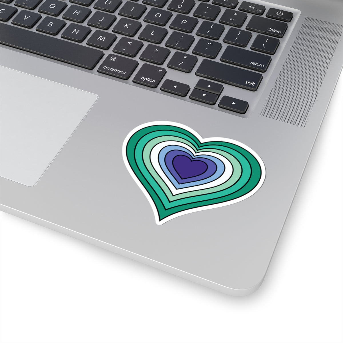 Close-up of the bottom right corner of a silver laptop featuring a 3 inch by 3 inch pride flag heart sticker in a Gay Men flag style applied to the surface.