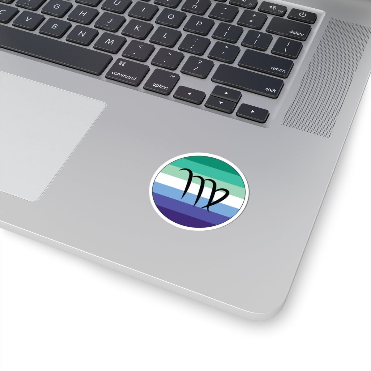 Close-up of the bottom right corner of a silver laptop featuring a 2 inch by 2 inch circular zodiac sticker with a centered Virgo glyph on a Gay Men pride flag style background applied to the surface.