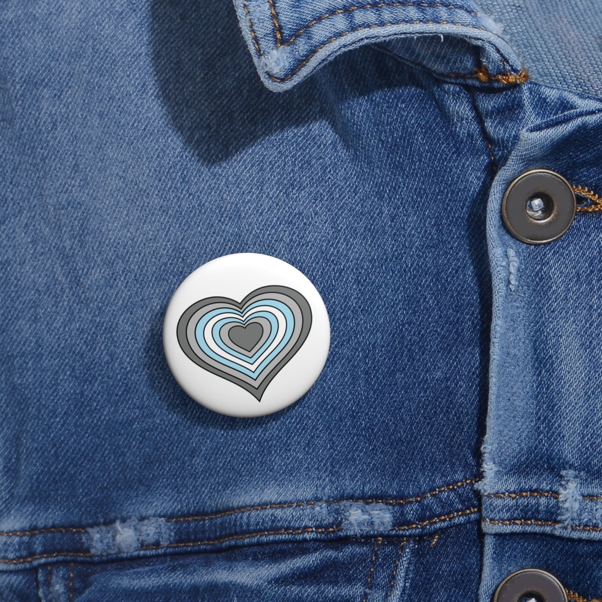 Close-up of a white circular pride button featuring a nested heart design inspired by the Demiboy pride flag in grays, white and blue, attached to a blue denim jacket at the right chest with the button clearly visible.