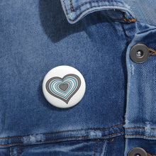 Close-up of a white circular pride button featuring a nested heart design inspired by the Demiboy pride flag in grays, white and blue, attached to a blue denim jacket at the right chest with the button clearly visible.