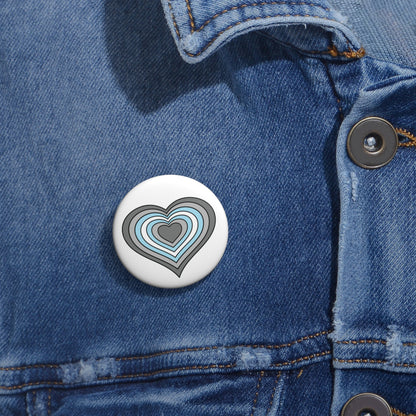 Close-up of a white circular pride button featuring a nested heart design inspired by the Demiboy pride flag in grays, white and blue, attached to a blue denim jacket at the right chest with the button clearly visible.