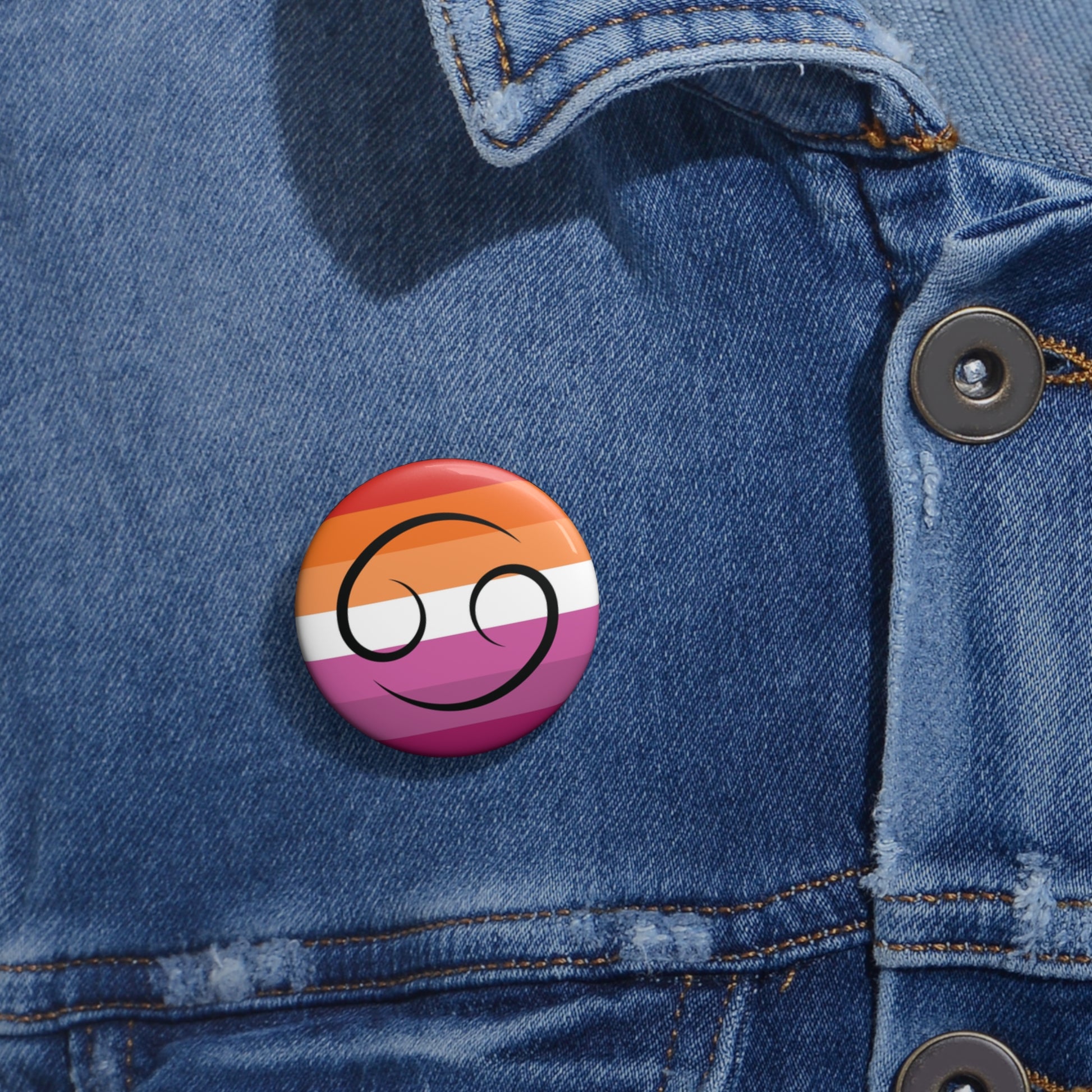 Close-up of a white circular pride button featuring the Cancer zodiac glyph centered over a lesbian pride flag background in oranges, white, and pinks attached to a blue denim jacket at the right chest with the button clearly visible.