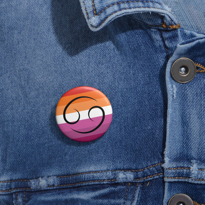Close-up of a white circular pride button featuring the Cancer zodiac glyph centered over a lesbian pride flag background in oranges, white, and pinks attached to a blue denim jacket at the right chest with the button clearly visible.