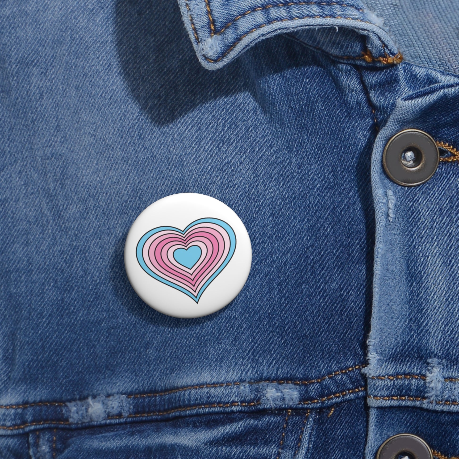 Close-up of a white circular pride button featuring a nested heart design inspired by the Transfem pride flag in blue and pinks, attached to a blue denim jacket at the right chest with the button clearly visible.
