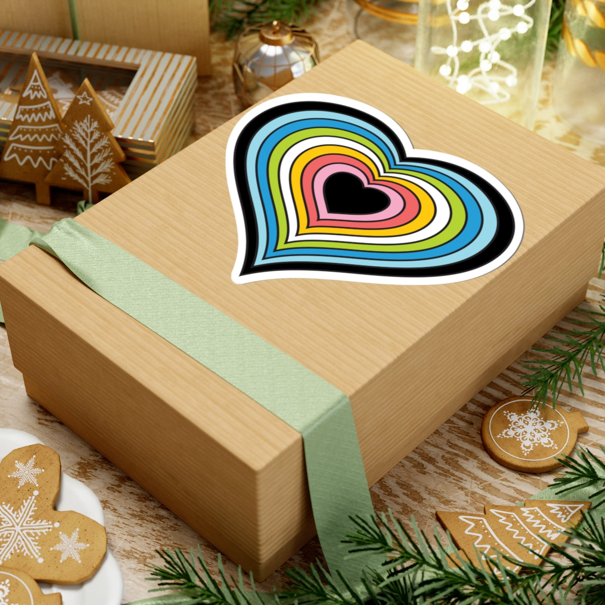 Gift box with a green ribbon on a festive background with cookies and Christmas decorations. Heart-shaped Queer pride sticker centered on the box.