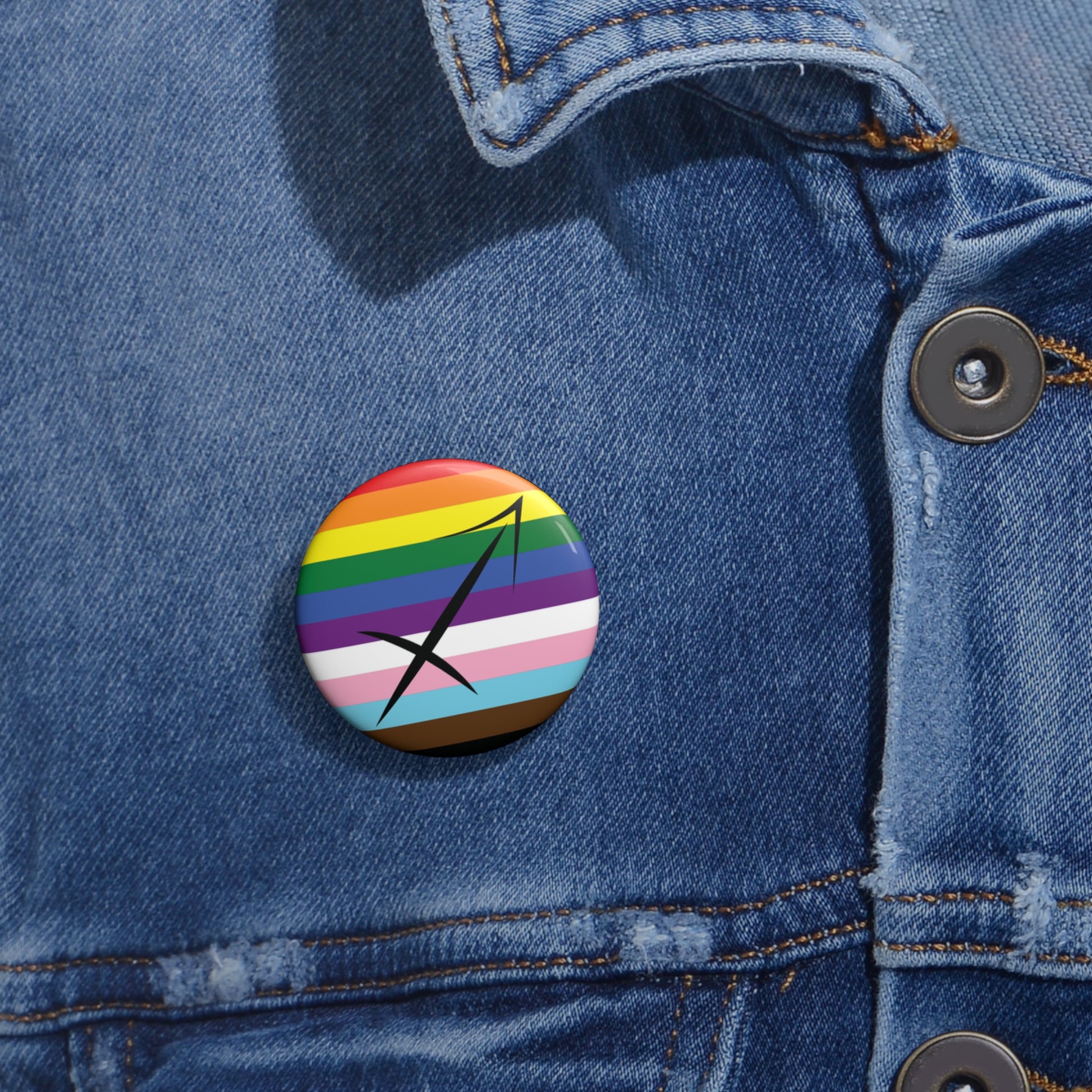 Close-up of a white circular pride button featuring the Sagittarius zodiac glyph centered over a Progress Pride flag background in rainbow stripes with black, brown, light blue, pink, and white elements, attached to a blue denim jacket at the right chest with the button clearly visible.