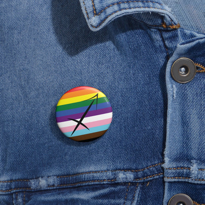 Close-up of a white circular pride button featuring the Sagittarius zodiac glyph centered over a Progress Pride flag background in rainbow stripes with black, brown, light blue, pink, and white elements, attached to a blue denim jacket at the right chest with the button clearly visible.