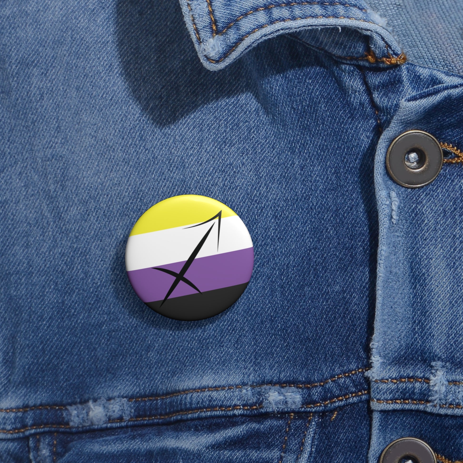 Close-up of a white circular pride button featuring the Sagittarius zodiac glyph centered over a nonbinary pride flag background in yellow, white, purple, and black, attached to a blue denim jacket at the right chest with the button clearly visible.