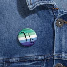 Close-up of a white circular pride button featuring the Capricorn zodiac glyph centered over a gay men (MLM) pride flag background in greens/teals, white, and blues, attached to a blue denim jacket at the right chest with the button clearly visible.