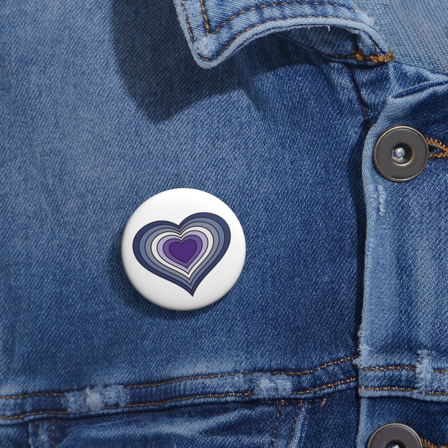 Close-up of a white circular pride button featuring a nested heart design inspired by the Butch pride flag in blues and purples, attached to a blue denim jacket at the right chest with the button clearly visible.