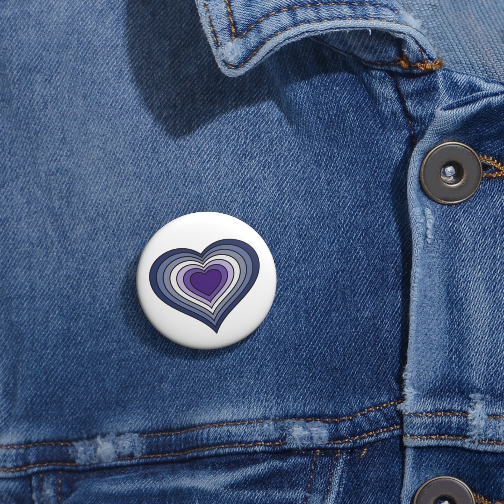 Close-up of a white circular pride button featuring a nested heart design inspired by the Butch pride flag in blues and purples, attached to a blue denim jacket at the right chest with the button clearly visible.