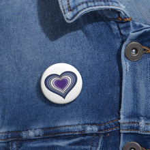 Close-up of a white circular pride button featuring a nested heart design inspired by the Butch pride flag in blues and purples, attached to a blue denim jacket at the right chest with the button clearly visible.