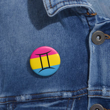 Close-up of a white circular pride button featuring the Gemini zodiac glyph centered over a Pansexual pride flag background in pink, yellow, and blue, attached to a blue denim jacket at the right chest with the button clearly visible.